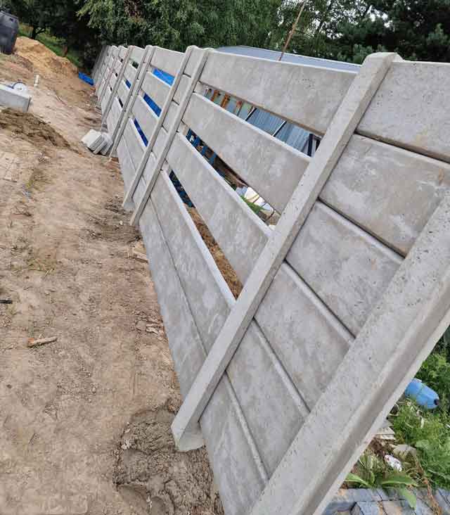 Ogrodzenie betonowe składające się z pionowych słupków i paneli, ustawione na trawniku, otoczone zielenią i drzewami w tle.
