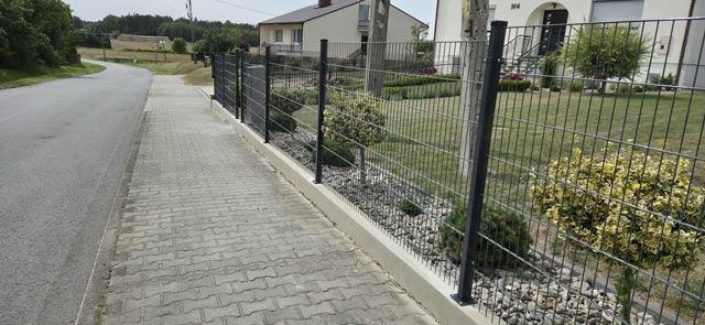 Metalowe ogrodzenie panelowe zamontowane na podmurówce wzdłuż chodnika przy posesji mieszkalnej.