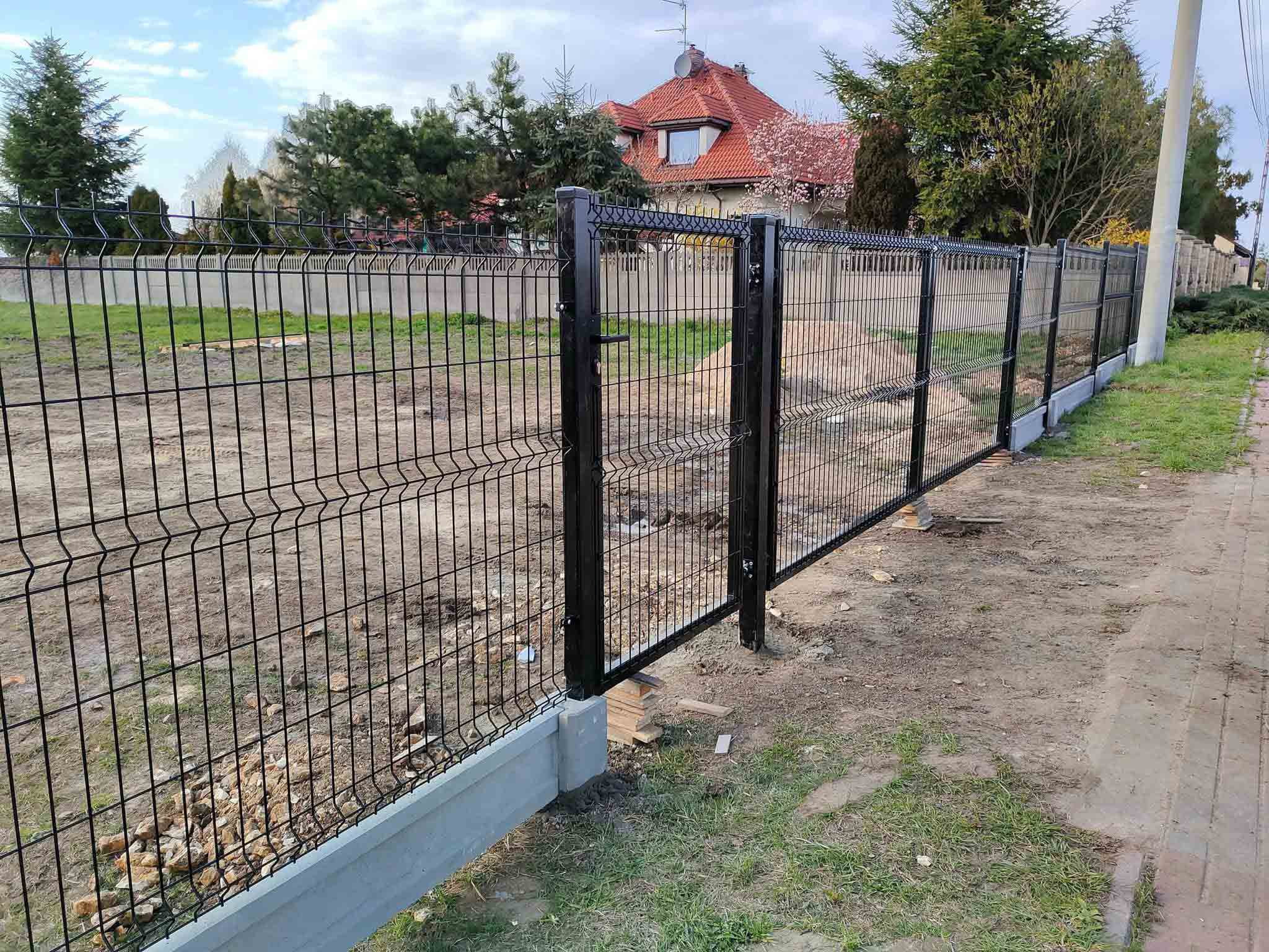 Czarne metalowe ogrodzenie panelowe na podmurówce wzdłuż chodnika przy nowoczesnym domu jednorodzinnym.