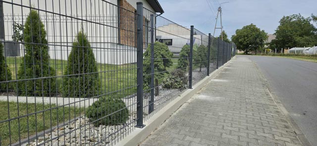 Grafitowe metalowe ogrodzenie panelowe wzdłuż chodnika przy zadbanej posesji z ozdobnymi krzewami.
