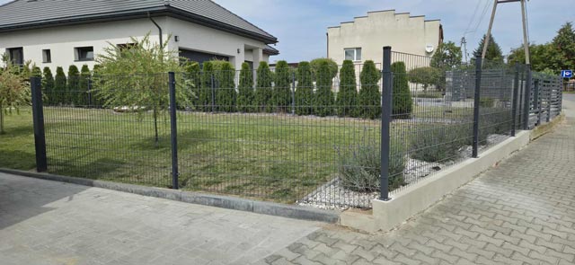 Metalowe ogrodzenie panelowe na podmurówce otaczające narożną posesję z trawnikiem i żywopłotem.