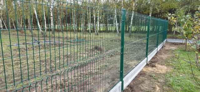 Zielone metalowe ogrodzenie panelowe na podmurówce wzdłuż działki z drzewami w tle.