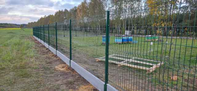 Zielone metalowe ogrodzenie panelowe na podmurówce biegnące wzdłuż rozległej działki przy otwartym terenie.