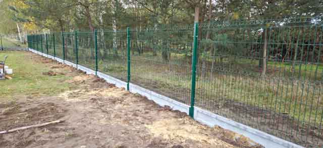 Zielone metalowe ogrodzenie panelowe na podmurówce wzdłuż zalesionej granicy działki.