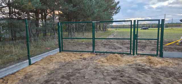 Zielone metalowe ogrodzenie panelowe z dwuskrzydłową bramą wjazdową, zamontowane na podmurówce przy otwartej działce.