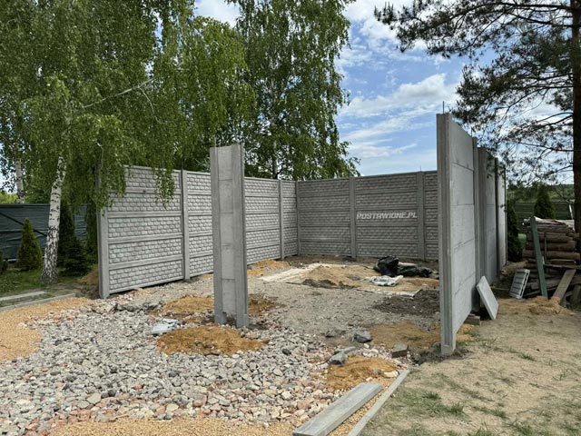 Montaż betonowego ogrodzenia z panelami o teksturze kamienia, na otwartej przestrzeni w słoneczny dzień.