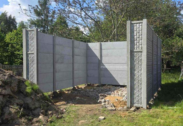 Betonowe ogrodzenie składające się z poziomych paneli, ustawione w kształcie kwadratu, z budynkami i lasem w tle.