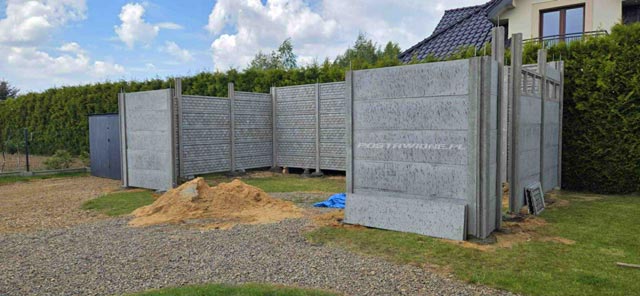 Montaż betonowego ogrodzenia składającego się z poziomych paneli, na otwartym terenie, otoczony lasem i budynkami w tle.