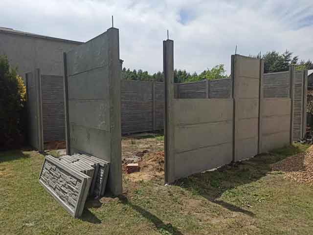 Montaż betonowego ogrodzenia składającego się z poziomych paneli, na otwartym terenie, otoczony lasem i budynkami w tle.