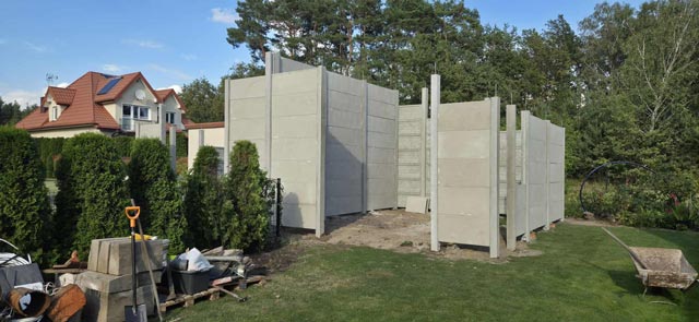 Montaż betonowego ogrodzenia składającego się z poziomych paneli, na otwartym terenie, otoczony lasem i budynkami w tle.