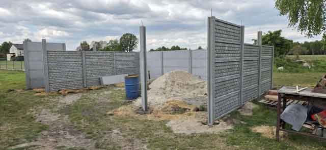 Montaż betonowego ogrodzenia składającego się z poziomych paneli, na otwartym terenie, otoczony lasem i budynkami w tle.