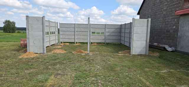 Montaż betonowego ogrodzenia składającego się z poziomych paneli, na otwartym terenie, otoczony lasem i budynkami w tle.